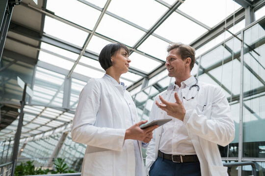Two doctors standing in atrium, discussing, holding digital tablet