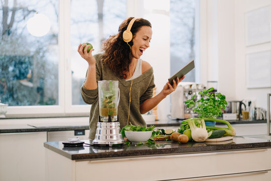 Fit woman standing in kitchen, preparing healthy smoothie, using online recipe