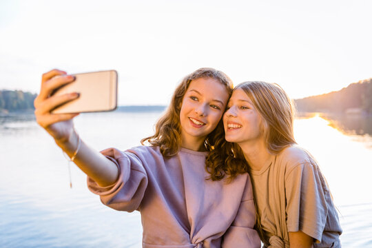 Smiling friends taking selfie on smart phone against lake