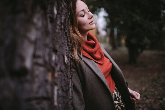 Portrait of woman with eyes closed leaning against tree trunk wearing turtleneck pullover