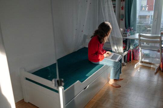 Girl with laptop sitting on bed in children's room looking out of window