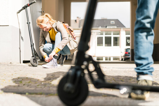 Young woman tying shoes, while friend is waiting with e-scooter