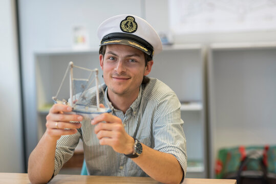 Portrait of smiling businessman sitting at desk in office wearing captain's hat and holding model ship