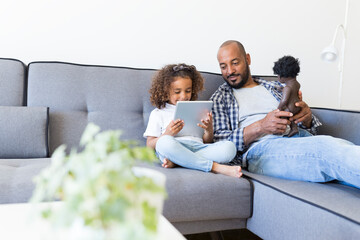 Father and daughter sitting on couch at home with doll and tablet