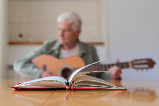 Close-up of diary and pen on table with senior man practicing guitar while sitting at home