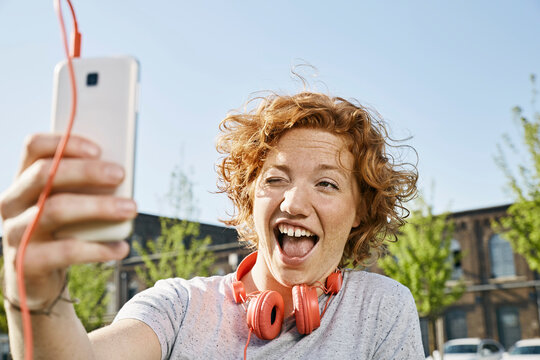 Playful young woman with headphones taking a selfie in urban surrounding