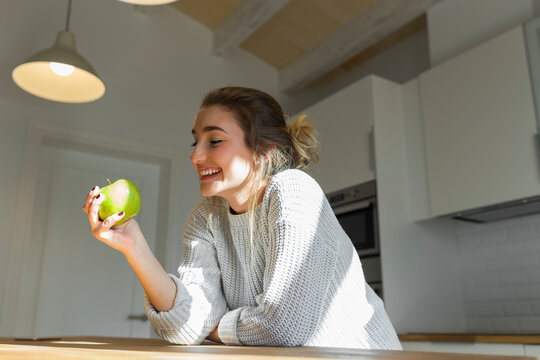 Young smiling woman holding green apple in the kitchen