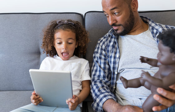 Father and excited daughter sitting on couch at home looking at tablet
