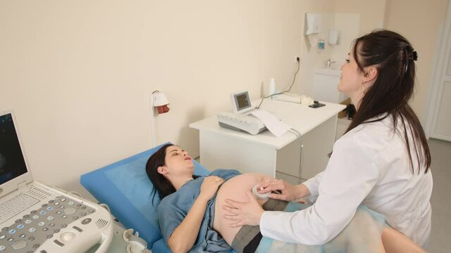 Pleasant pregnant woman with various sensors and belts on her belly lying on couch while female therapist monitoring heartbeat of baby. Professional health care for brunette in expectation.