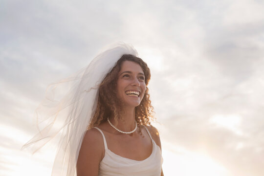 Cheerful young bride looking away while standing at beach against sky during sunset
