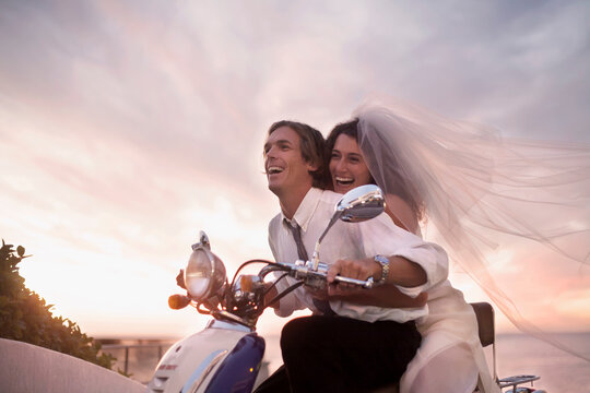 Cheerful newlywed couple in wedding dress riding motor scooter during sunset