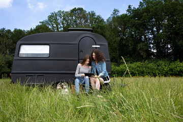 Two best friends sitting on a meadow in front of black caravan using digital tablet