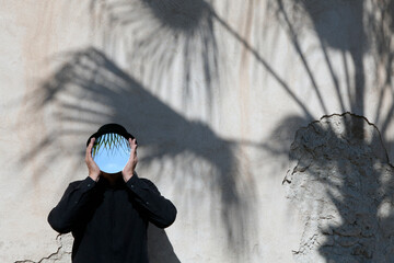 Morocco, Essaouira, man wearing a bowler hat holding mirror in front of his face at a wall