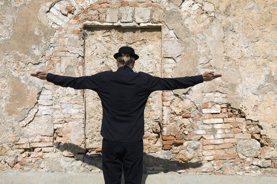 Rear view of man wearing a bowler hat standing at stone wall with arms outstretched