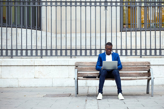 Young businessman wearing blue suit jacket sitting on bench and using laptop