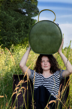Portrait of young woman sitting in suitcase on a meadow with hatbox on top of her head