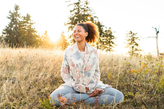 Cheerful woman sitting with crossed leg on grass while doing yoga during sunset