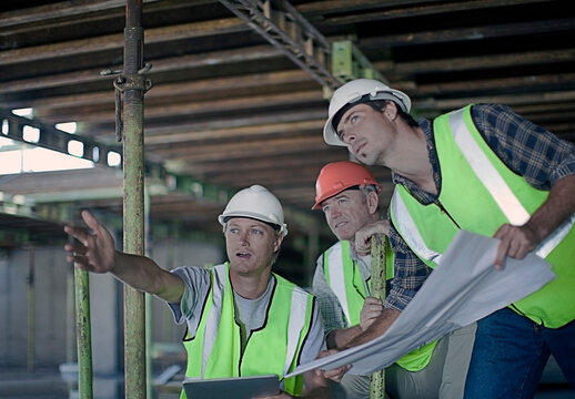 Male construction worker pointing while discussing with coworkers over blueprint at construction site