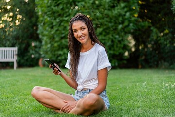 Smiling braided woman with mobile phone sitting cross legged in garden