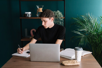 Handsome young man writing in book while sitting with laptop at table
