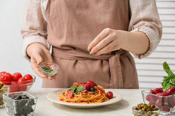 Woman preparing tasty Pasta Puttanesca at table in kitchen, closeup