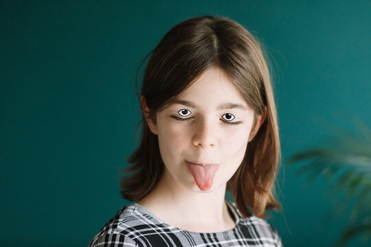 Close-up of girl with googly eyes and make-up sticking out tongue against wall at home