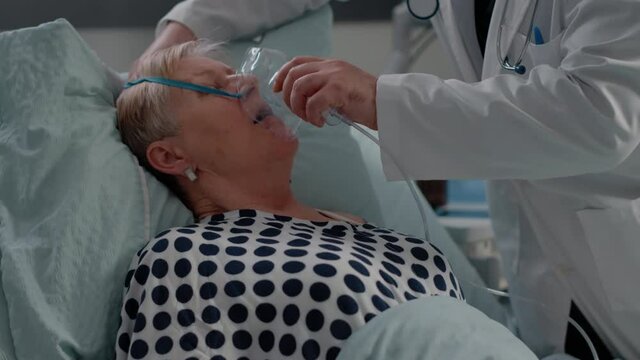Doctor and nurse giving assistance to heavy breathing woman, using oxygen tube to cure respiratory issues in bed. Medical team helping aged patient with hyperventilating illness.