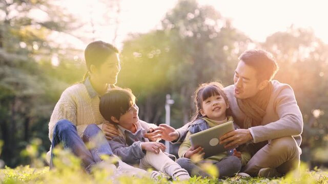 asian family with two children sitting on ground relaxing outdoors in park at dusk