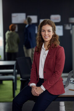 Portrait of smiling businesswoman sitting on desk in office with colleagues in background