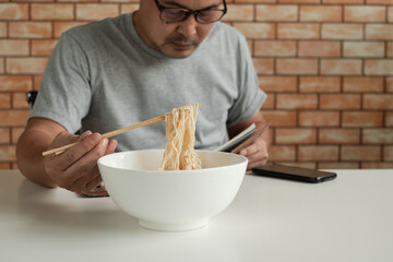 Asian male worker reads an appointment book while eating instant noodles in white bowl with chopsticks on table in brick wall background office during a lunchtime break, a hastily unhealthy lifestyle.