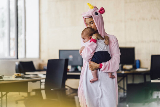 Young mother wearing unicorn onesie, standing in office, holding her son in her arms