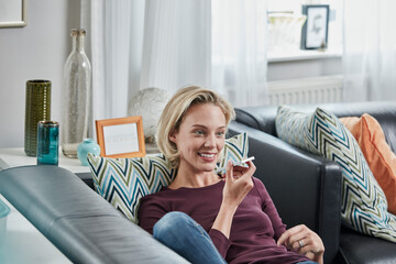 Smiling young woman using cell phone lying on couch at home