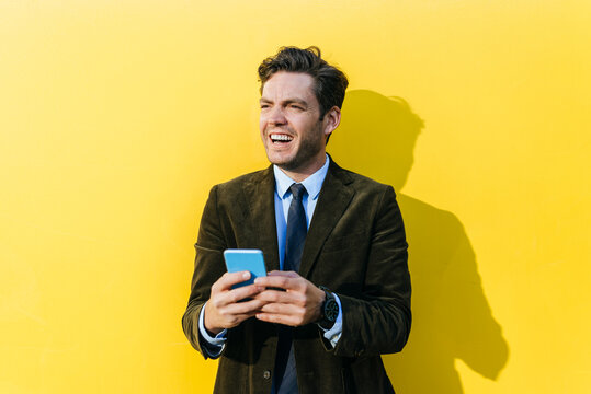 Happy businessman with smartphone in front of yellow wall