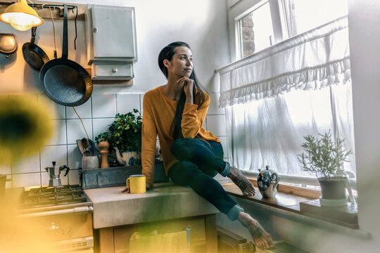Young woman sitting on kitchen counter at home looking out of window