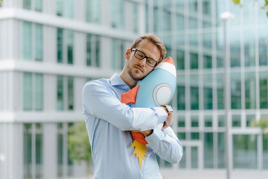 Young businessman standing in front of modern office building, hugging toy rocket