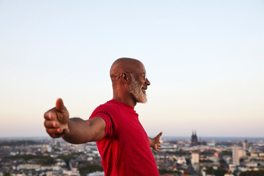 Happy completely bald man standing with arms outstretched on building terrace in city during sunset