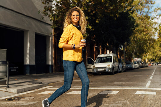 Happy Young Woman Crossing Street While Walking With Disposable Cup In City On Sunny Day