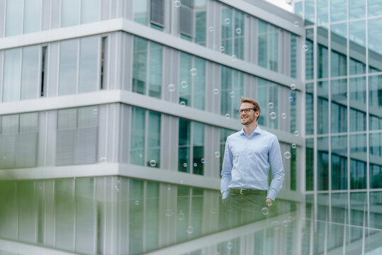Young businessman standing in front of modern office building, watching soap bubbles
