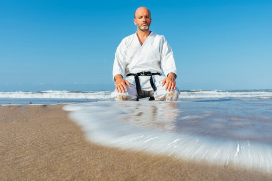 Mature man meditating while kneeling at beach against clear blue sky