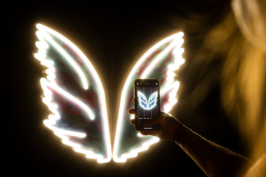 Arm of young woman taking smart phone photo of white glowing neon wings