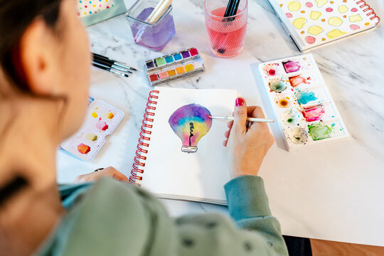 Woman painting light bulb with watercolors on spiral notebook