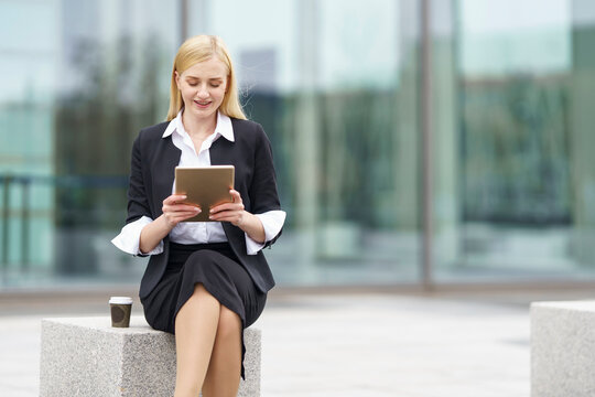 Blond businesswoman using digital tablet while sitting on bench against building