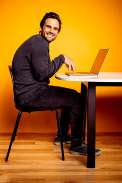 Smiling handsome man using laptop while sitting at table against yellow background