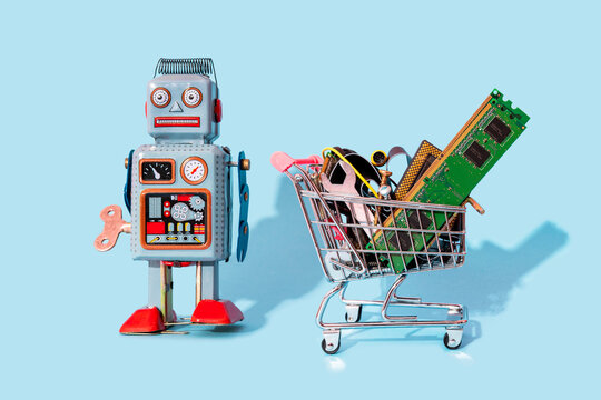Studio shot of vintage robot toy standing by miniature shopping cart filled with electronic equipment