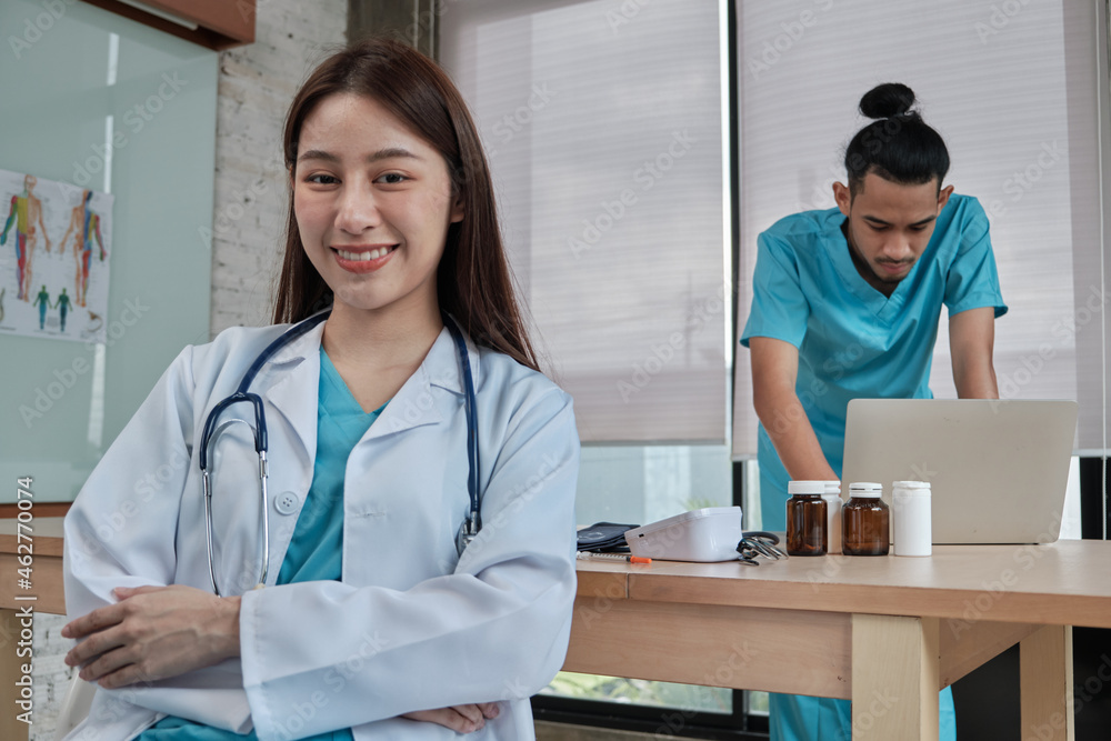 Wall mural Portrait of beautiful female doctor of Asian ethnicity in uniform with stethoscope. Smile and looking at camera in a hospital clinic, male partner working behind her, two professional persons.