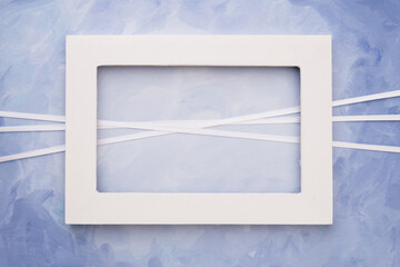 Empty simple white picture frame on a blue painted canvas with ribbon decoration; Simple template for a greeting card
