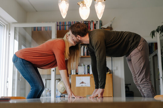 Couple standing on chairs, kissing, leaning on dining table