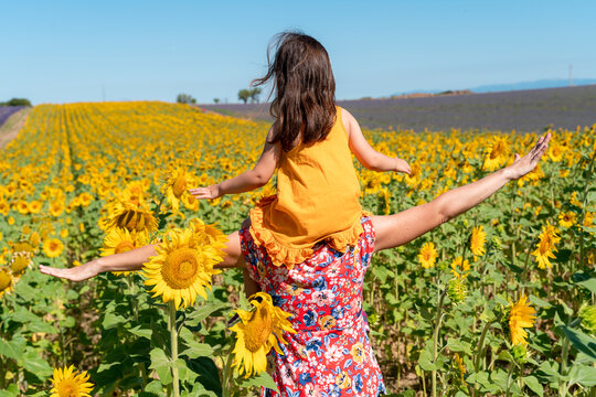 Mother carrying daughter on shoulders in sunflower field during summer