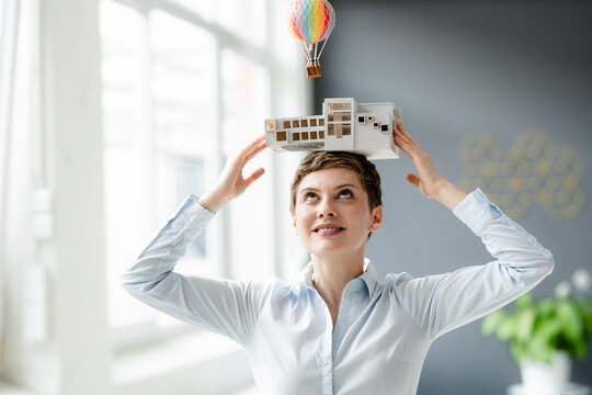 Smiling businesswoman carrying architectural model on her head with hot-air balloon floating above