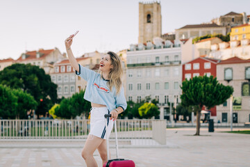 Cheerful woman taking selfie with mobile phone while standing with suitcase on footpath in city
